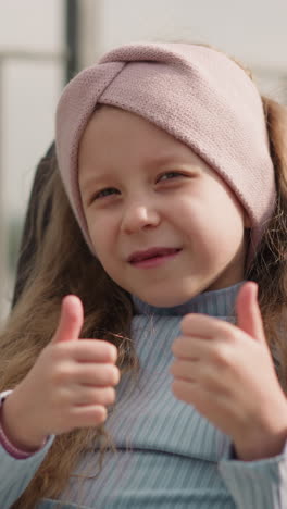 cute toothless girl with sad eyes smiles in camera showing thumbs up gesture against blurred fencing. redhead child with disability sits in wheelchair closeup