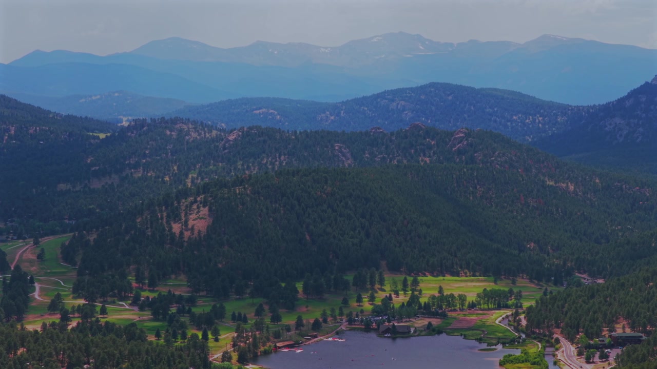 Veduta aerea estiva con drone della Evergreen Lake House di Evergreen, Colorado, con il Monte Blue Sky, le colline della Front Range delle Montagne Rocciose, il paesaggio del campo da golf di Denver coperto di neve, cielo velato e nuvole soleggiate, con movimento circolare a destra.