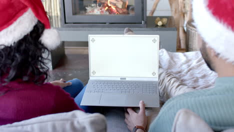 Biracial-couple-wearing-santa-claus-hats-using-laptop-with-copy-space-on-screen-at-home,-slow-motion