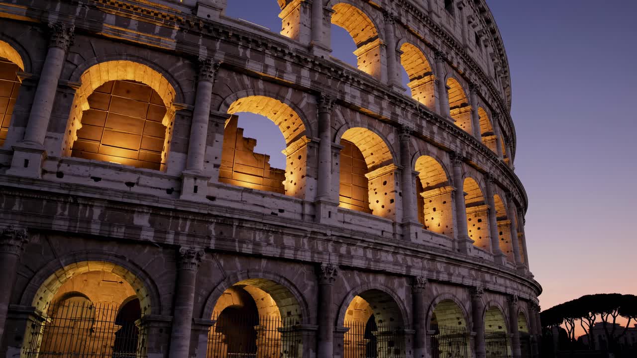 Tomada de vídeo em ângulo baixo do Coliseu ao entardecer, destacando seus arcos iluminados.