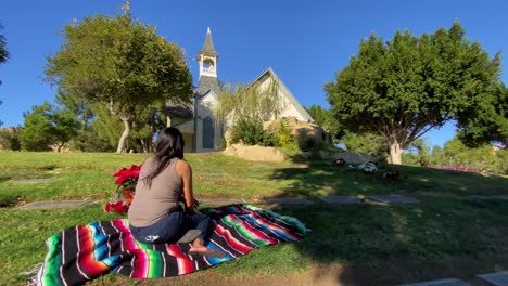Rear-view-female-sitting-on-colourful-striped-blanket-paying-respect-praying-in-front-of-church-on-sunny-day