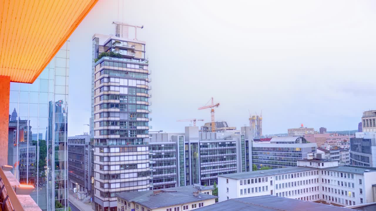 View of a uniquely designed skyscraper under construction, standing out in the city skyline, adorned by green balconies, under an overcast sky, framed by an orange awning