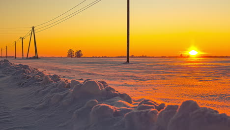 Tiro-De-Lapso-De-Tiempo-Del-Campo-De-Invierno-Cubierto-De-Nieve-Con-Torre-De-Transmisión-Durante-La-Puesta-De-Sol-Dorada-En-El-Horizonte