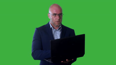 un hombre calvo escribiendo y programando una computadora portátil y mirando atentamente a la pantalla mientras está de pie contra un fondo de pantalla verde