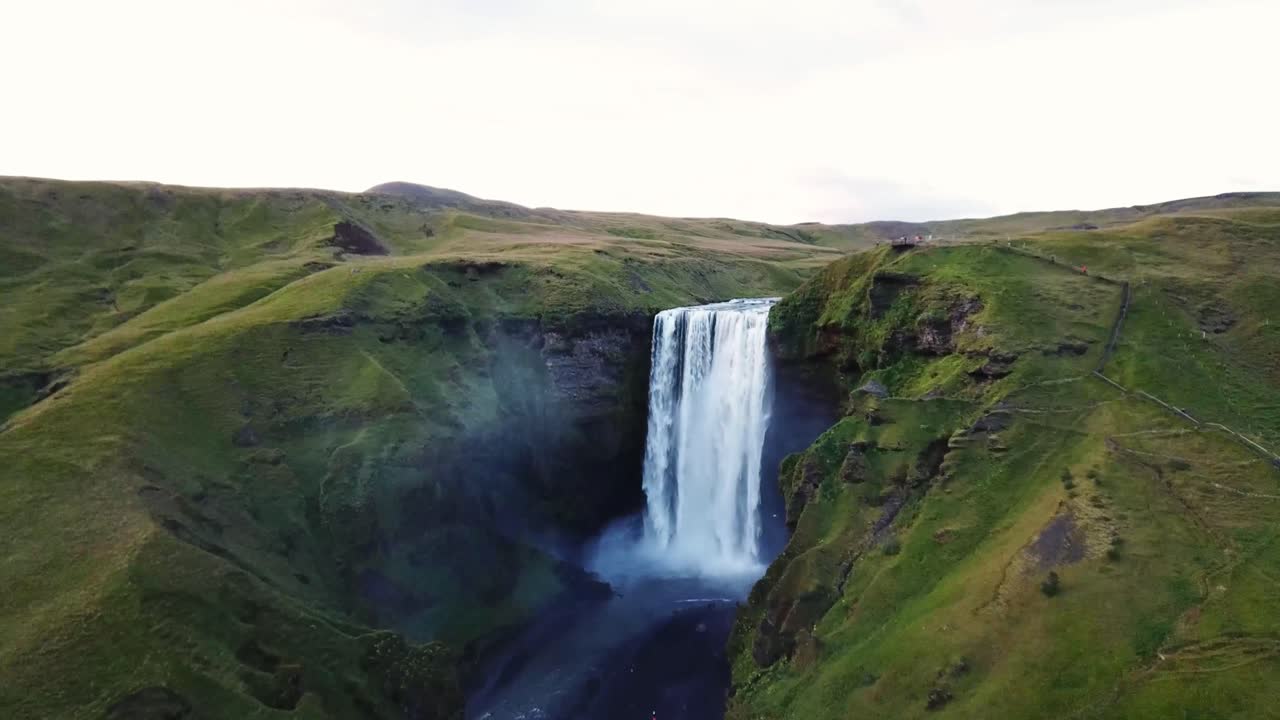 Powerful Skógafoss waterfall plunging into a rocky gorge with a river flowing through a green valley in Iceland, creating a breathtaking natural landscape, drone dolly in establishing shot