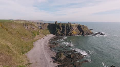 Impresionante-Panorama-Del-Castillo-De-Dunnottar-En-Escocia-Con-El-Océano,-Olas-Rompiendo-Y-Campos-De-Color-Verde-Amarillo-En-El-Fondo