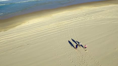 Descending-aerial-shot-of-beachgoers-and-fishermen-walking-slowly-up-the-beach-amid-car-tracks-and-footprints,-casting-long-dramatic-shadows-in-the-afternoon-sun