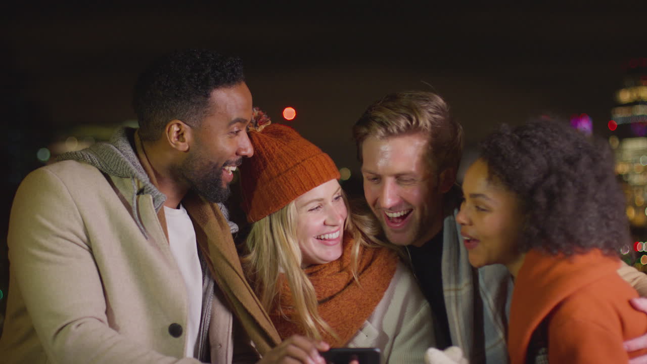 Group of friends wearing winter coats and scarves posing for selfie on mobile phone with city lights in background - shot in slow motion