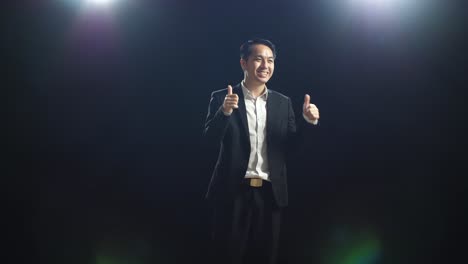 smiling asian speaker man in business suit showing thumbs up gesture and looking around while standing in the black screen studio