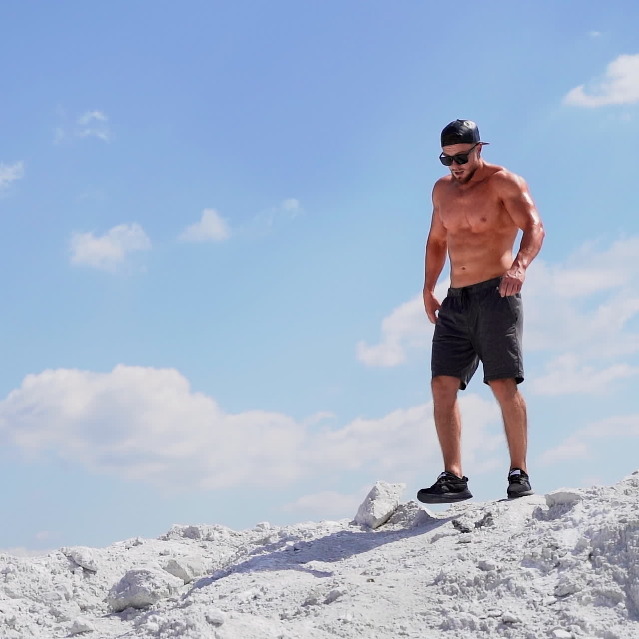 Muscular man walking on white canyon. Strong shirtless man in black glasses going down in a slow motion on mountain under clear sky.