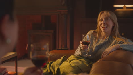 two female friends relaxing and laughing on sofa in lounge with cosy fire holding glass of wine