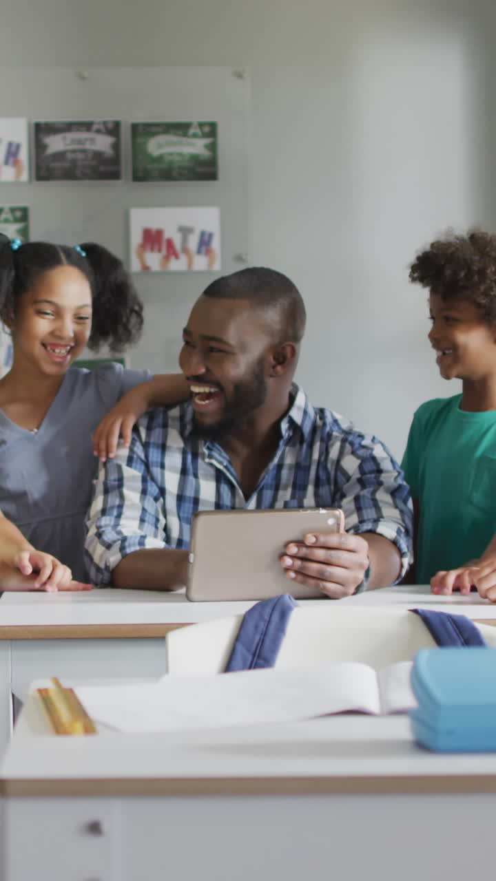 video de un feliz maestro afroamericano y una clase de alumnos diversos trabajando en una tableta