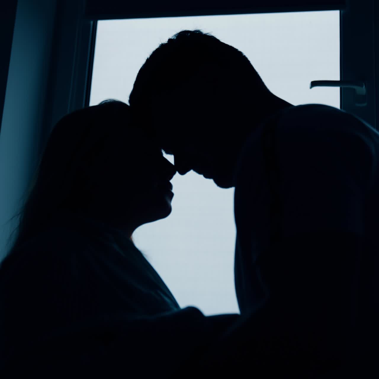 Silhouettes of expecting couple standing at the window. Man strokes big wife's belly and then embraces her. Low angle view