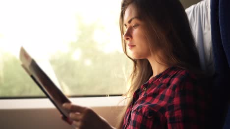 young, pretty girl travelling by modern train. sitting next to the window and reading in plaid shirt. side view. blurred window in motion