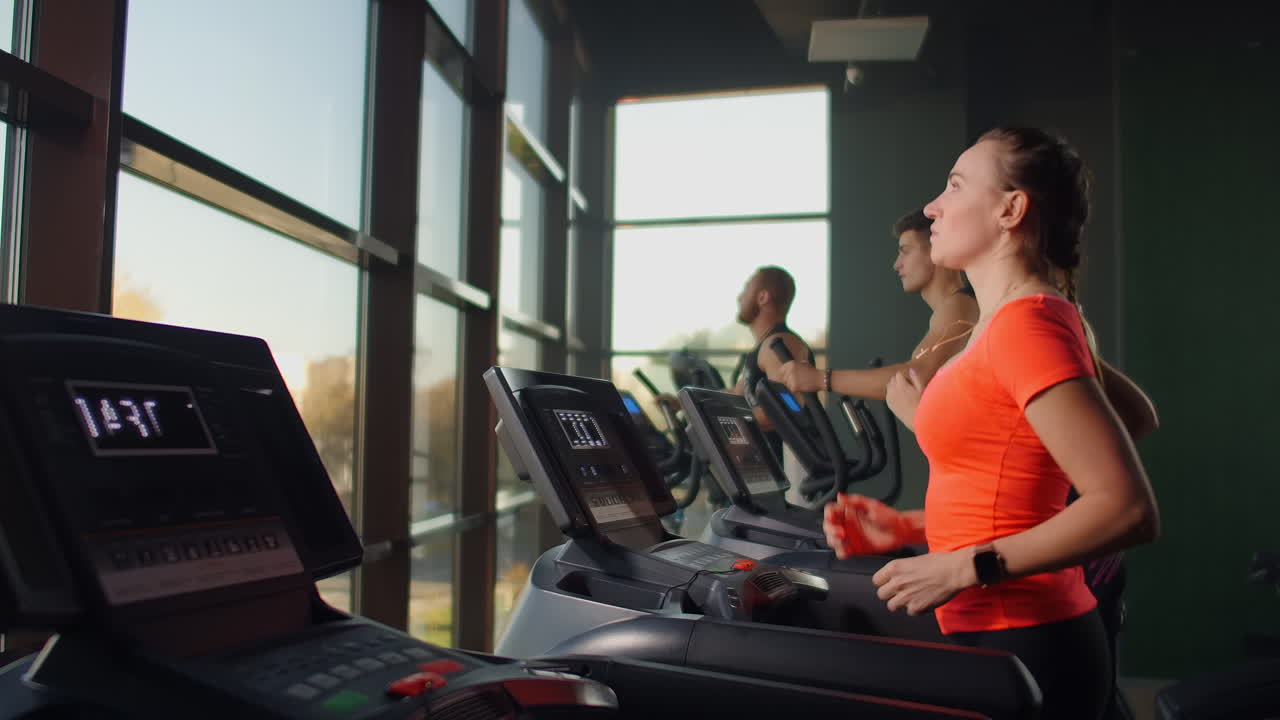A young beautiful woman and three people running on a treadmill in a fitness room performing a cardio workout. Indoor running warm-up before training in slow motion.