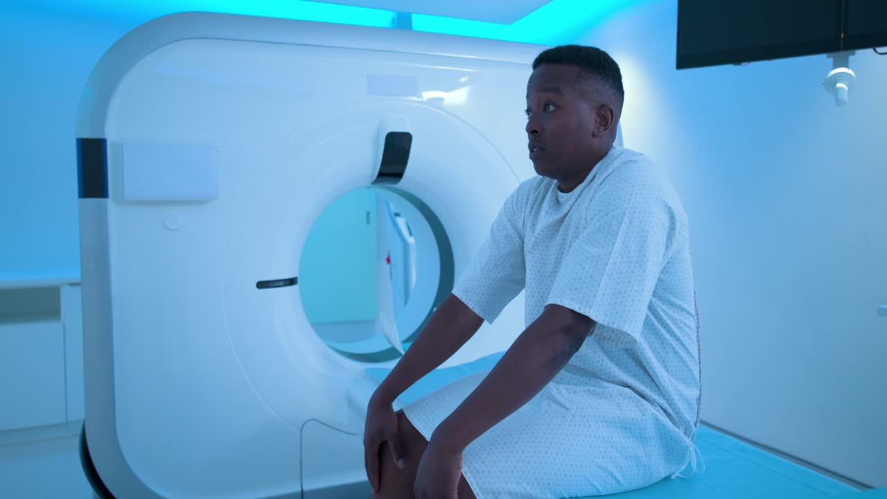 Uomo africano adulto seduto su un lettino da visita, in camice, che si copre e scopre il viso mentre attende una scansione.