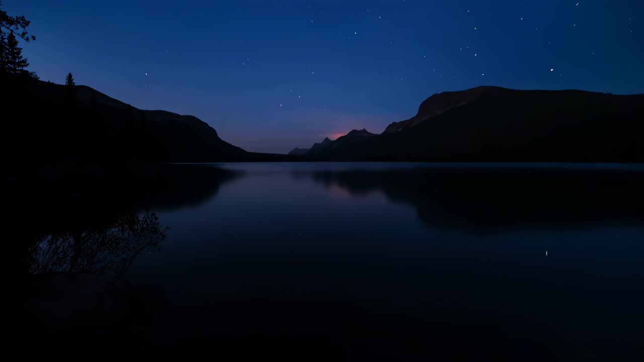 Scure creste montuose circondano un lago calmo sotto un cielo sereno pieno di stelle, con un debole bagliore dell'orizzonte che si riflette sull'acqua immobile