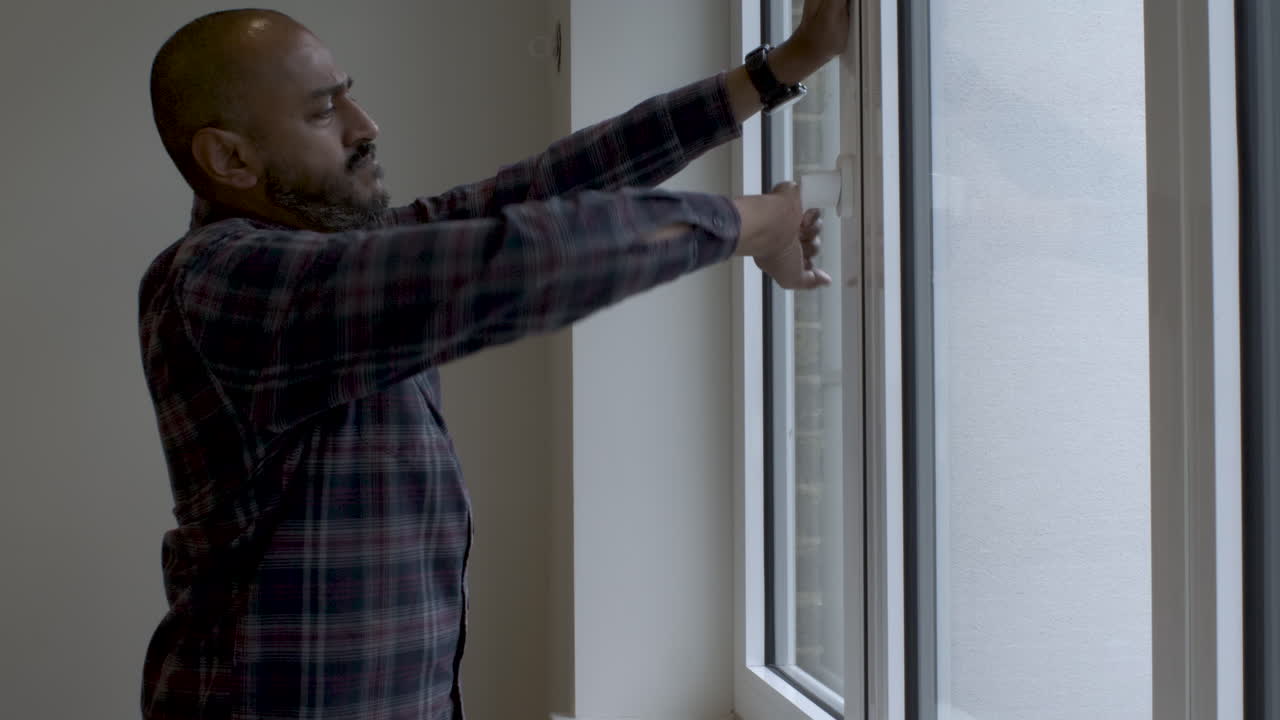 Indian Bald Male In Casual Attire Walking Towards Window To Close It By Turing Handle And Checking It
