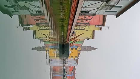 Water-surface-pov-view-of-Burano-colorful-houses-and-canal-with-moored-boats-and-belltower-in-background,-Italy