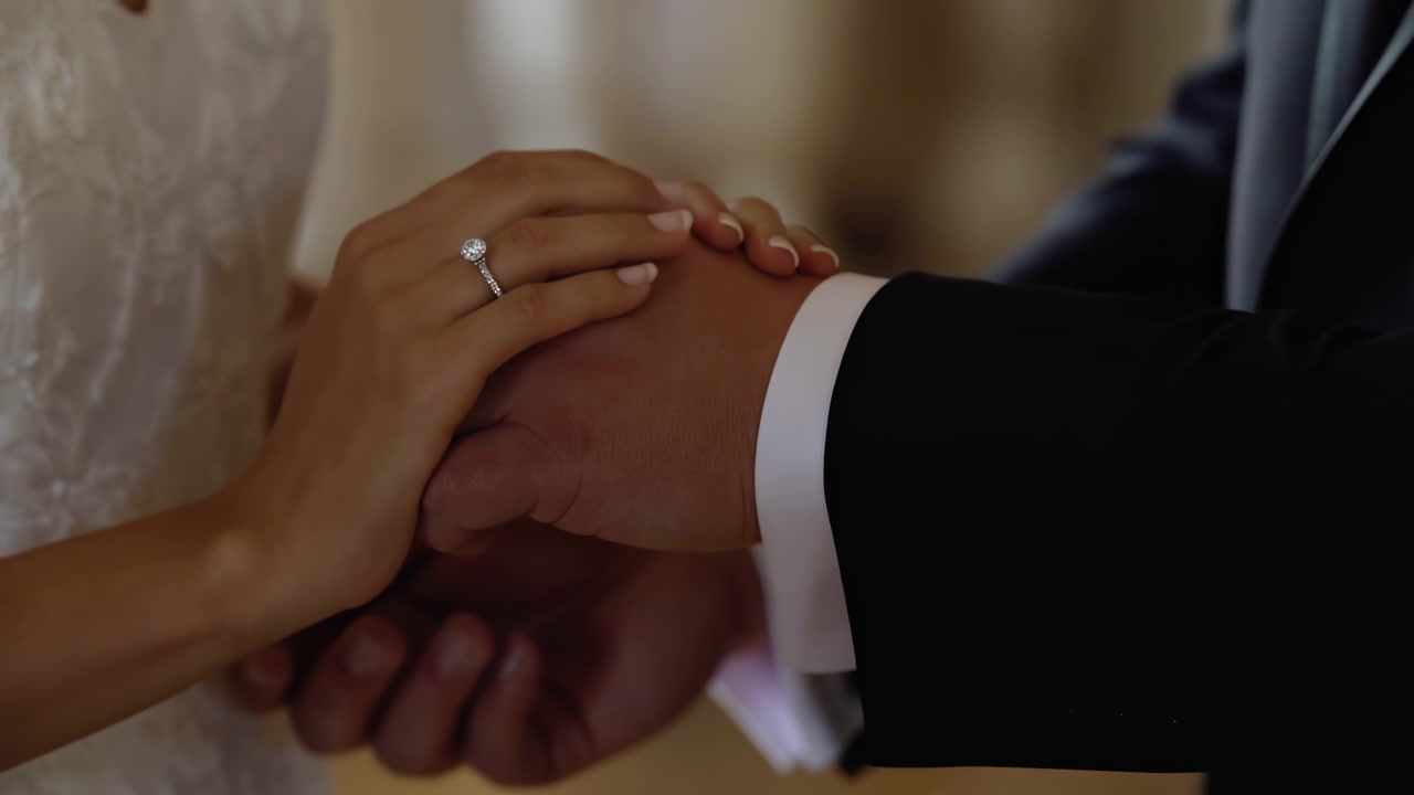 Close Up Bride And Groom Hands Gently Clasped Over Suit Sleeve And Lace Gown, Engagement Ring Sparkling, Warm