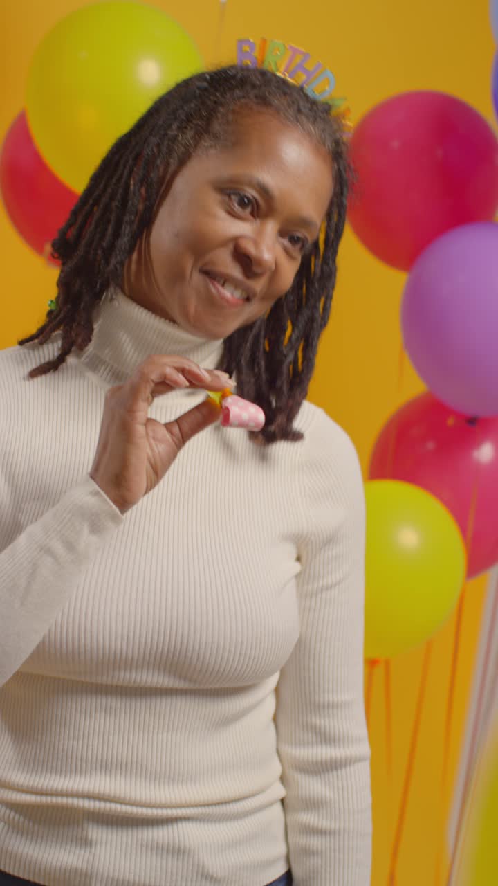Vertical Video Studio Portrait Of Woman Wearing Birthday Headband Celebrating With Balloons And Party Blower