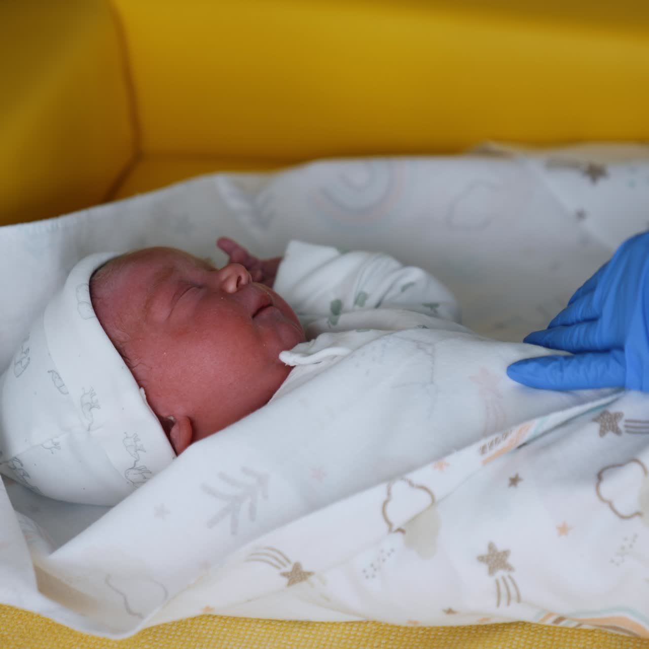 Neonato che indossa una cuffietta e una tutina bianca. Un medico con i guanti avvolge il bambino in una coperta. Primo piano