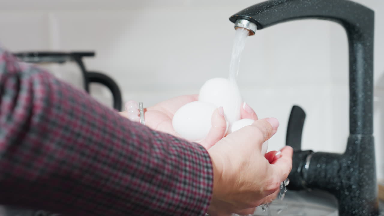 primer plano de una mujer lavando a mano tres huevos blancos bajo un grifo en una cocina moderna, lleva un traje a cuadros y un brazalete de cuentas, haciendo hincapié en la higiene