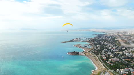 Drone-follow-Powered-paraglider-flyover-turquoise-water-by-Glyfada-Coastline,-Greece