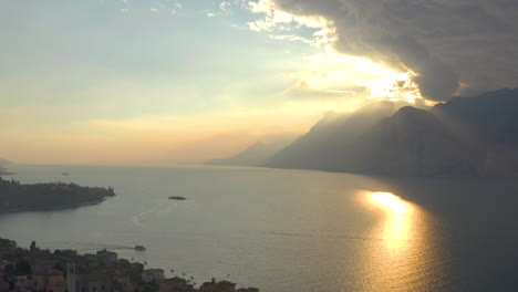 Increíble-Toma-Aérea-Del-Lago-Garda-Al-Atardecer-Con-Nubes-Dramáticas-Y-Luz-Dorada-En-El-Fondo