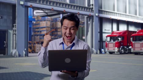 asian male professional worker standing with his laptop, outside of logistics distributions warehouse, he raises his fist up with screaming goal