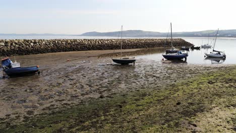 Aerial-view-boats-in-shimmering-low-tide-sunny-warm-Rhos-on-Sea-rock-jetty-seaside-sandy-beach-marina-orbit-left-low