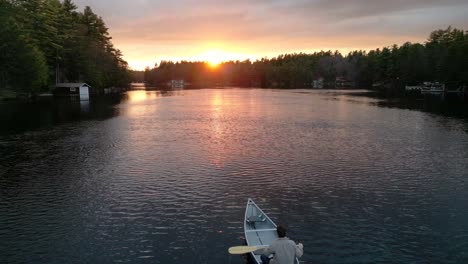 Person-canoeing-on-a-tranquil-lake-during-sunset,-surrounded-by-trees-and-reflecting-the-golden-light-on-the-water