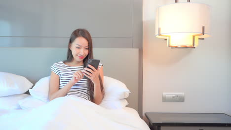 beautiful happy asian woman typing first morning message on smartphone from bed