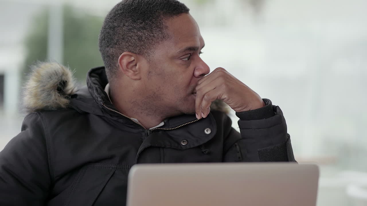 Afro-American middle-aged man in black jacket with fur hood working on laptop outside
