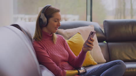 woman with prosthetic arm wearing wireless headphones watching film on  mobile phone on sofa