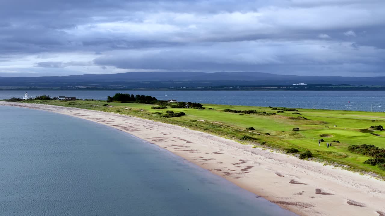 Drone glides above sandy beach, green fields, and seaside village under dramatic cloudy skies