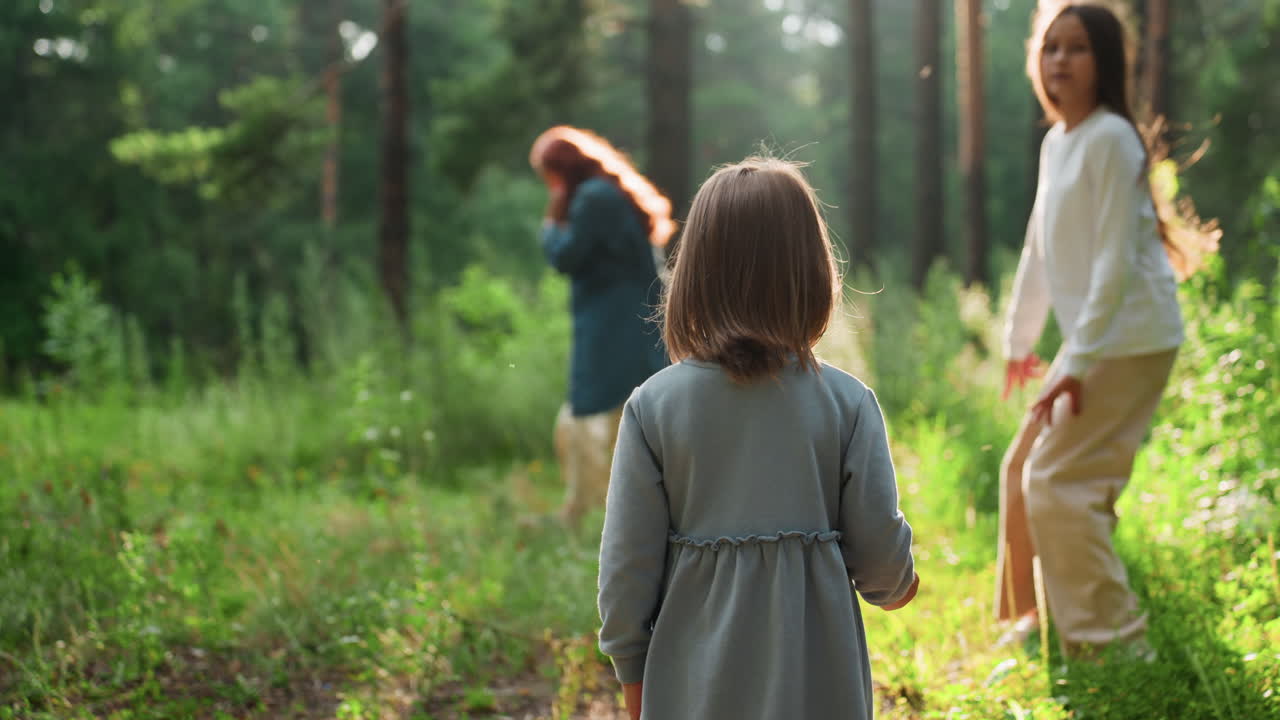 Back view of child standing still on forest path watching sister run toward mother who is picking flowers in warm sunlight, creating tender family moment filled with connection, and natural beauty