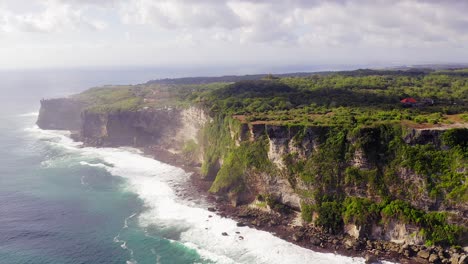 uluwatu cliffs, pull back aerial view revealing the sharp edge cliff with lush green landscape, tertiary limestone layers and indian ocean waves breaking on the beach, bali, indonesia