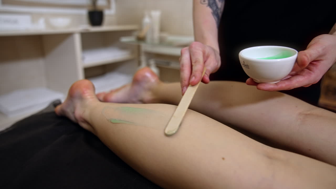 Hands apply light green cream with wooden paddle on the female leg. Masseur spreads the cream by the client's leg.
