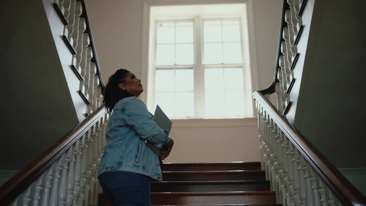 Kvinde i denimjakke holder en bærbar computer og begynder at gå op ad trætrapper mod et vindue for at nyde udsigten.