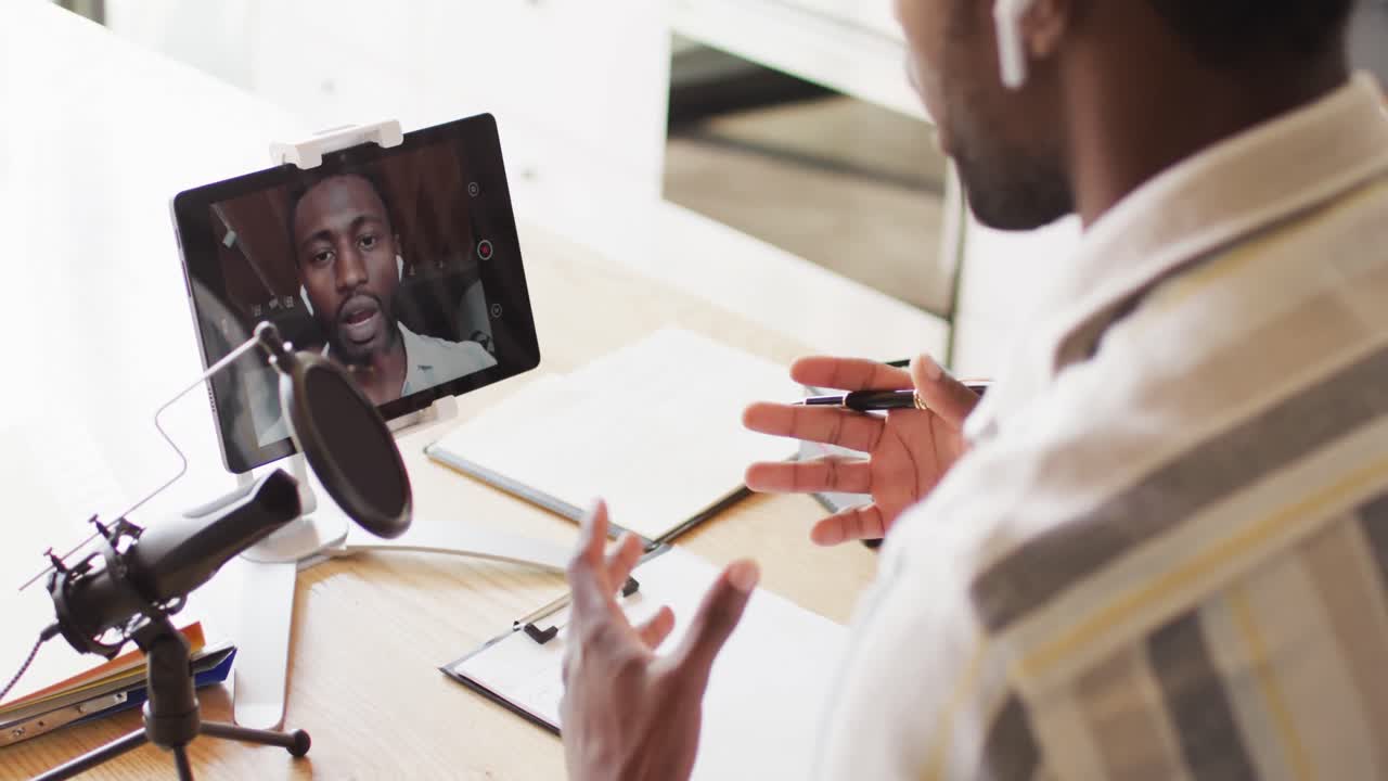 Happy african american man sitting at table in kitchen, using tablet and making vlog