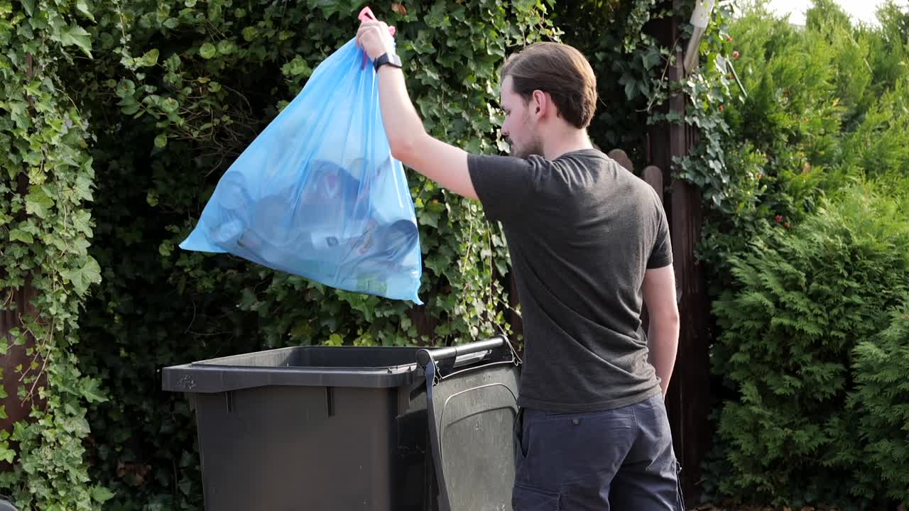 Caucasian male taking out trash in plastic bag to bin in garden