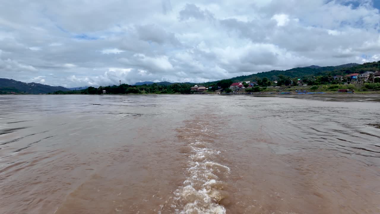 A view of the Mekong River from a slow boat in Houayxay Laos featuring brown water a cloudy sky and a lush green mountain landscape