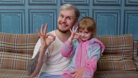 Father-and-little-child-daughter-kid-in-pajamas-sit-on-couch-in-room-smiling-showing-ok-gesture-sign