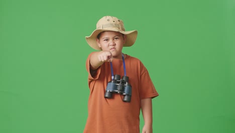asian tourist boy with a hat and binoculars pointing his finger towards you and furiously speaking against blaming you on the green screen background. boy researcher, travel tourism adventure concept