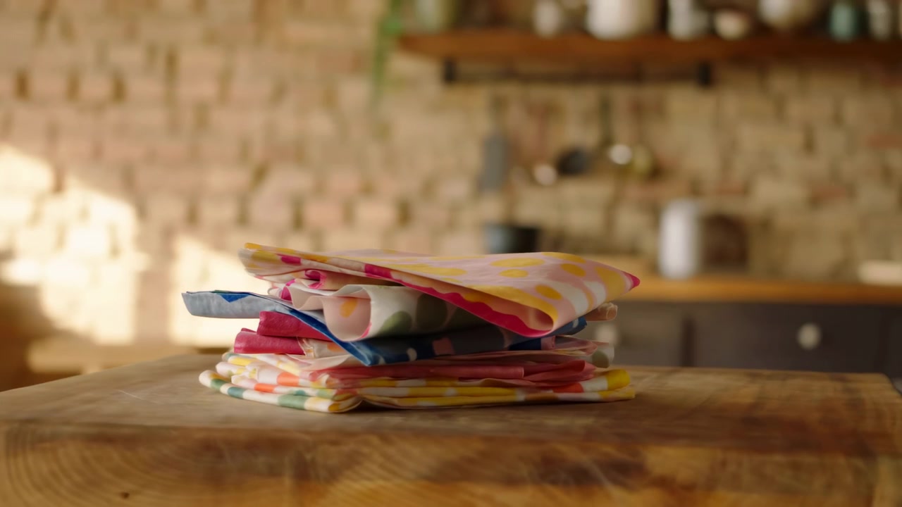 Layered patterned textiles remain centered on the rustic worktop while the background moves across the frame, with golden daylight, warm wood tones, open shelving, and shallow focus.