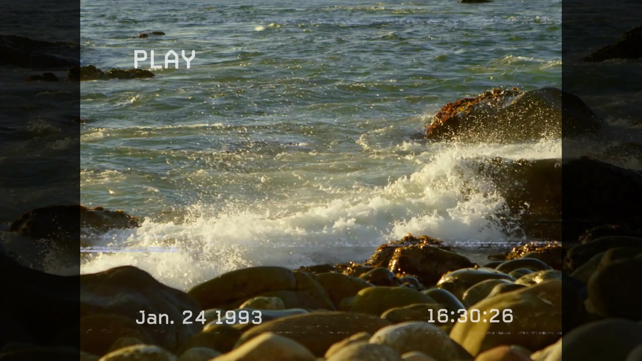 Wave pushing right rock, spraying foam over boulders, receding, rewetting, showing PLAY timestamp