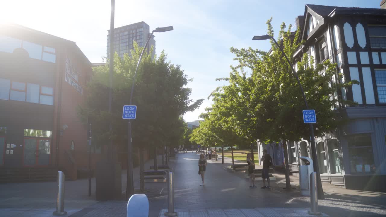 Handheld sideways shot of people standing in pedestrianised area in Sheffield city centre, England. Buildings and trees in background.