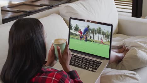 compuesto de una mujer sentada en casa sosteniendo un café viendo un partido de hockey en una computadora portátil