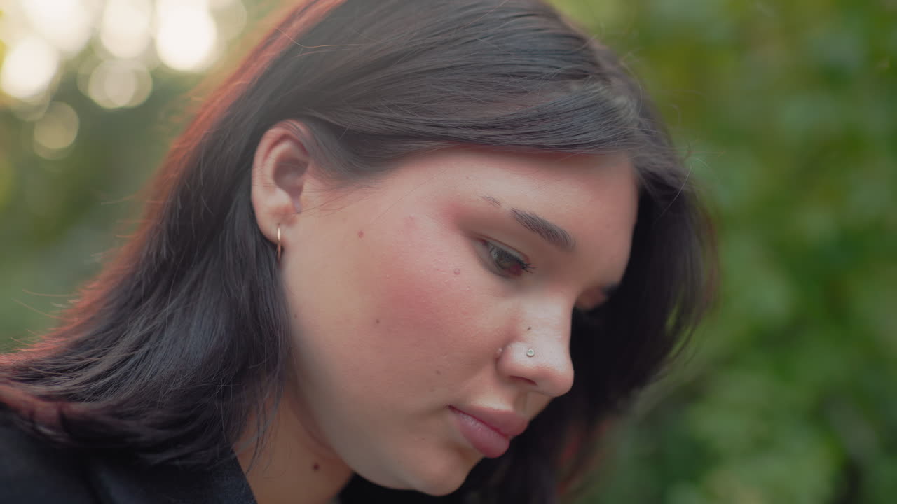 primer plano lateral de una chica de piel clara leyendo en un parque, con la mano acomodándose el pelo al lado de la oreja, suave fondo bokeh de un bosque, enfoque tranquilo, luz suave de otoño, momento sereno al aire libre, ambiente de estudio tranquilo